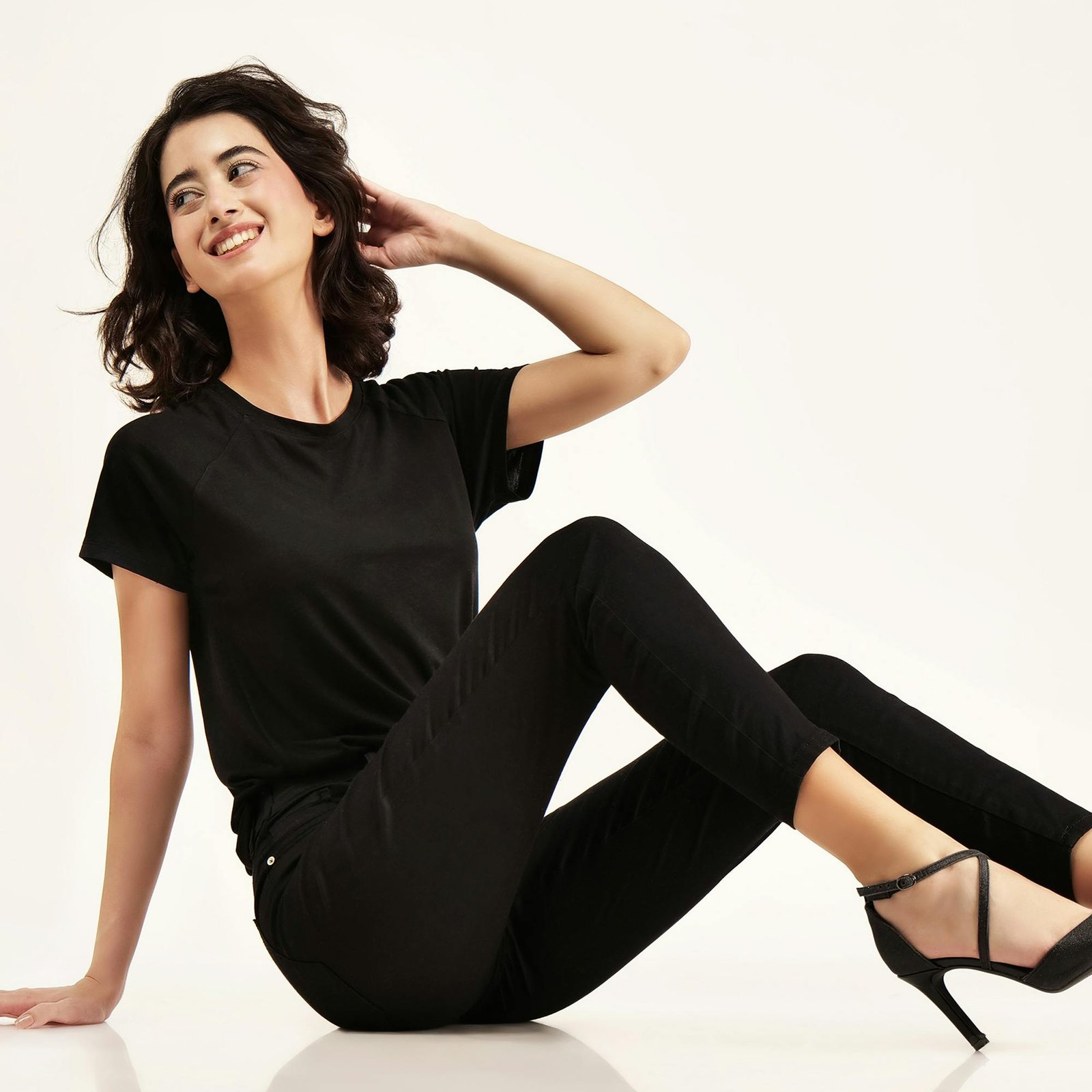 Smiling female model in black attire sitting against a white backdrop, showcasing casual fashion.